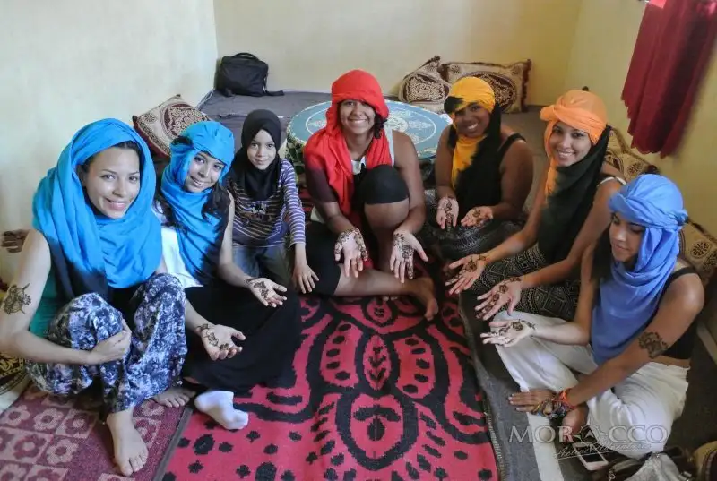educational tours in Morocco: A group of young women and a girl in colourful headscarves sitting on a red carpet in a Berber family home, showing henna designs on their hands