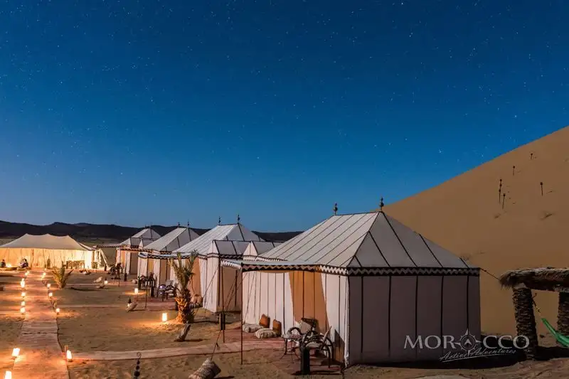 Morocco honeymoon: Row of white luxury tents lit by lanterns beside a sand dune under a starry night sky