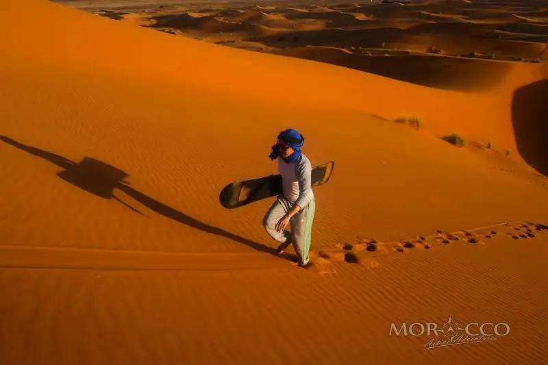 Sandboarding in Morocco: a rider slides down an Erg Chebbi dune in Merzouga, with two quads parked on the crest